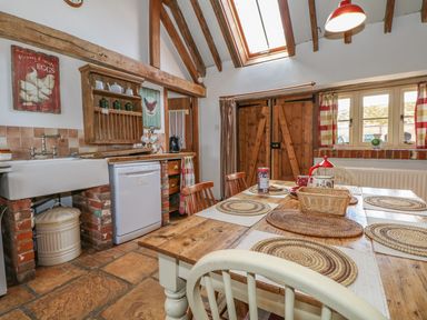 A kitchen with a wooden dining table set with placemats and a basket next to a farmhouse sink and wooden cabinets at Downs View Dairy in West Tytherley