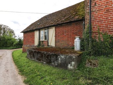 An exterior view of a red brick building with moss on the roof and a metal milk can on a raised concrete platform at Downs View Dairy West Tytherley