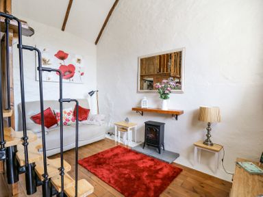 A living room with a spiral staircase a beige couch with red pillows a red rug a small black stove and a wall mirror at Rose Cottage in Penrhiwllan