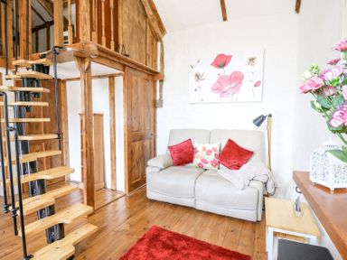 A small living room with a beige couch red cushions a wooden spiral staircase and floral wall art at Rose Cottage in Penrhiwllan