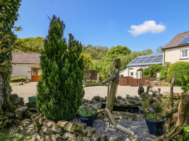 A garden area with a wooden human figure sculpture near potted plants and a pond with buildings in the background at Rose Cottage in Penrhiwllan