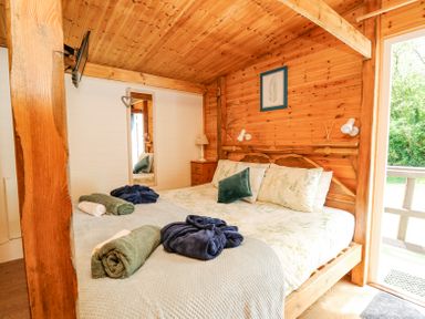 A bedroom with a wooden ceiling and walls a bed with floral bedding rolled towels robes a wall mirror and a bedside table with a lamp at Brook Lodge in Penrhiwllan