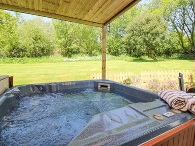 A bubbling hot tub under a wooden canopy with towels on the edge overlooking a grassy fenced garden at Brook Lodge in Penrhiwllan