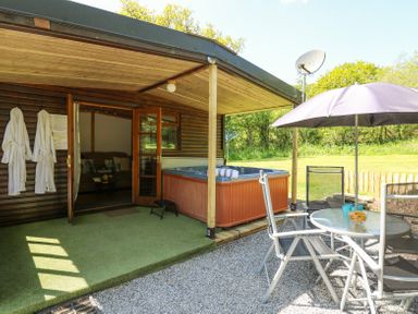 An outdoor patio with a hot tub two robes hanging on the wall a glass table with chairs and an umbrella at Brook Lodge in Penrhiwllan