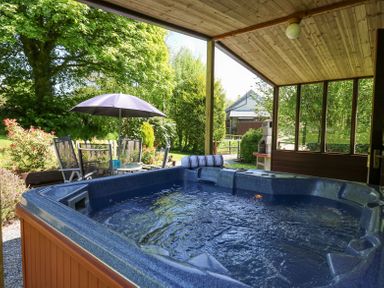 A covered patio area with a hot tub and a garden table with umbrella outside at Brook Lodge in Penrhiwllan