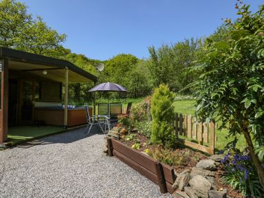 An outdoor patio area with a wooden shed a hot tub garden furniture with an umbrella and a small garden with wooden fencing at Brook Lodge in Penrhiwllan