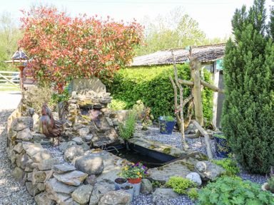 An outdoor garden area with a stone pond, waterfall, potted plants, metal rooster sculpture, and wooden figure at Brook Lodge in Penrhiwllan
