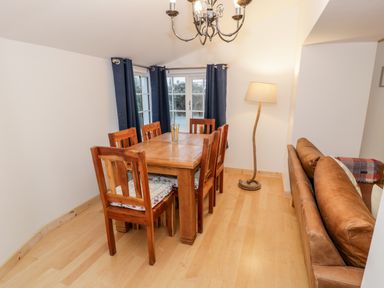 A dining area with wooden table and chairs next to a window with dark blue curtains and a floor lamp beside a brown leather sofa at Pentowyn in Holyhead