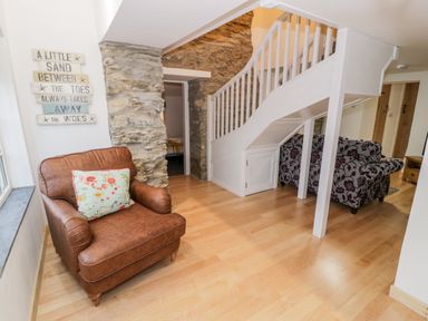 An interior room with a brown armchair, a floral patterned sofa, wooden flooring, white staircase, and stone wall at Pentowyn in Holyhead