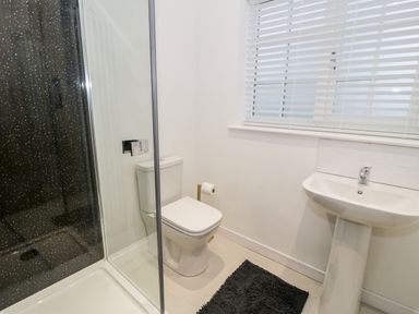 A bathroom with a shower enclosure a toilet a sink with a faucet a window with blinds and a black floor mat at Pentowyn in Holyhead