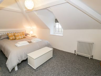 A bedroom with a bed featuring gray bedding and yellow pillows a window with a gray blind a radiator a bedside table with a lamp and a white storage chest at Pentowyn in Holyhead
