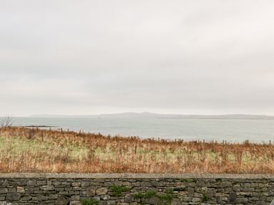 A stone wall with grass and dry plants in front of a large body of water and hills in the distance at Pentowyn in Holyhead