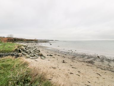 A sandy beach with rocks and grass next to a calm sea under a cloudy sky at Pentowyn in Holyhead