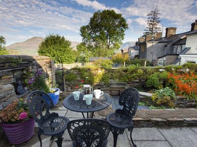 An outdoor patio area with a metal table and three chairs with cups and a French press surrounded by plants and stone walls at Moelwyn View Cottage in Llan Ffestiniog
