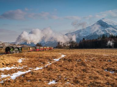 A steam train moving through a grassy field with patches of snow and mountains in the background at Moelwyn View Cottage in Llan Ffestiniog