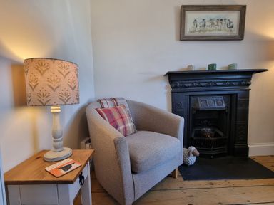 A corner with a beige armchair next to a fireplace and a side table with a lamp and a book at Moelwyn View Cottage in Llan Ffestiniog
