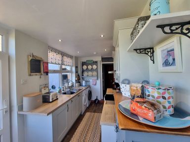 A narrow kitchen with wooden countertops white cabinets appliances and patterned window blind at Moelwyn View Cottage in Llan Ffestiniog