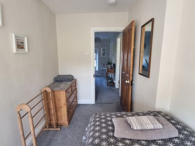 A bedroom with a bed covered in patterned bedding and towels a wooden dresser with a folded blanket and a clothes drying rack next to it and an open door showing a hallway at Moelwyn View Cottage in Llan Ffestiniog