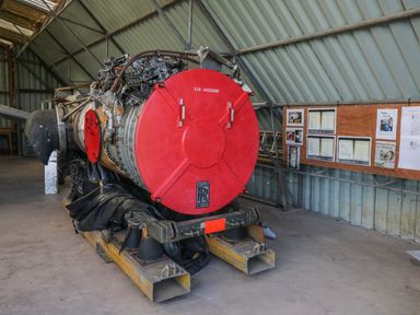 A Rolls Royce jet engine with a red cover inside a metal hangar with informational boards on the wall at Swan Lodge Castle Farm in Wedmore