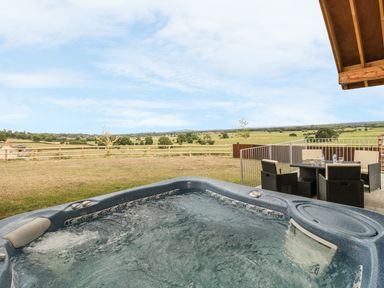 An outdoor hot tub with bubbling water next to a patio dining set overlooking a fenced grassy field at Swan Lodge Castle Farm in Wedmore