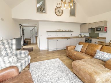 A living room with brown leather sofa, plaid armchair, white rug, and an open kitchen with wooden countertops at Swan Lodge Castle Farm in Wedmore