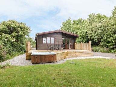 A wooden cabin with a deck table and chairs and a hot tub in a grassy area surrounded by trees at Oak Lodge Constantine near Falmouth