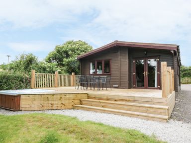 A wooden cabin with a deck and outdoor table and chairs near greenery at Oak Lodge in Constantine near Falmouth
