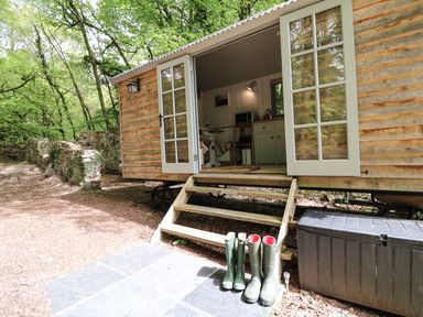 A wooden hut with open double doors showing an interior with kitchen cabinets and a table in a wooded area with boots on the steps at Rock View Shepherd's Hut in Gulworthy near Tavistock