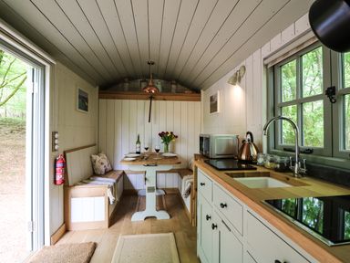 A kitchen and dining area with wooden countertops a sink induction cooktop a dining table with wine glasses and a bottle and built in benches at Rock View Shepherd's Hut in Gulworthy near Tavistock