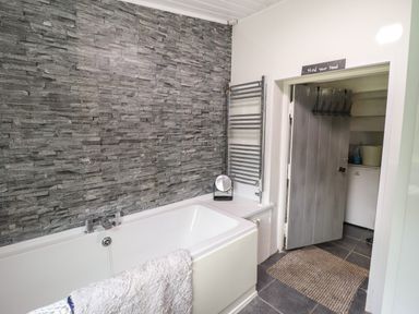 A bathroom with a bathtub beside a stone textured wall a towel hanging on the tub and an open door leading to another room at Gwenallt in Malltraeth