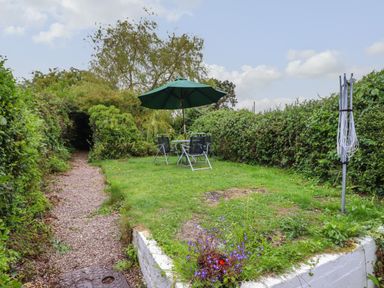 A garden area with a patio table and four chairs under a green umbrella and a rotary clothesline surrounded by hedges at Gwenallt in Malltraeth