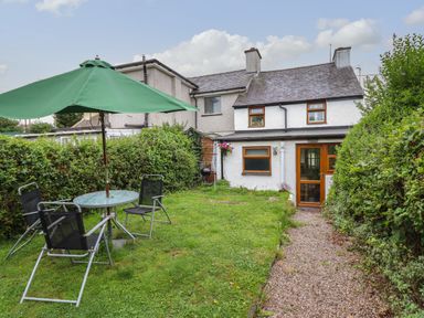 A backyard garden with a green umbrella and black chairs around a glass table next to a path leading to a white house with wooden framed windows at Gwenallt in Malltraeth