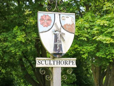 A village sign with a shield showing a windmill a flower and an eagle above the name Sculthorpe with trees in the background