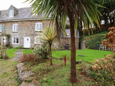 A stone house with white windows and door surrounded by a garden with palm trees and green grass at Lanvean Farmhouse Lanvean near St Mawgan