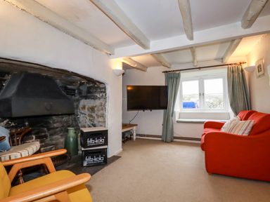 A living room with a stone fireplace wooden chairs a red sofa a wall-mounted television and a window with curtains at Lanvean Farmhouse Lanvean near St Mawgan