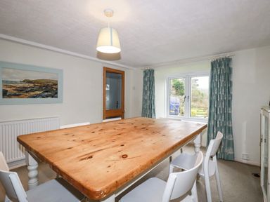 A dining room with a large wooden table white chairs a window with blue curtains and a painting on the wall at Lanvean Farmhouse in Lanvean near St Mawgan