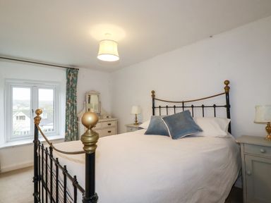 A bedroom with a metal bed frame white bedding two blue pillows bedside tables with lamps and a window with floral curtains at Lanvean Farmhouse Lanvean near St Mawgan