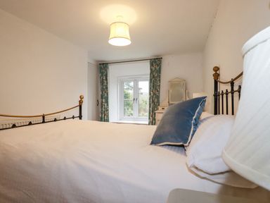 A bedroom with a metal bed frame white bedding blue pillow a window with floral curtains and a dresser at Lanvean Farmhouse in Lanvean near St Mawgan