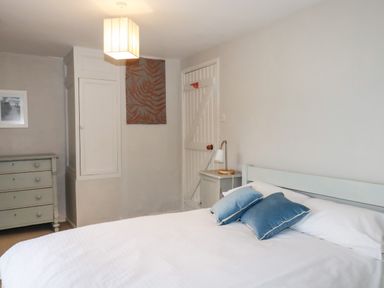 A bedroom with a bed with white sheets and blue pillows a bedside table with a lamp a chest of drawers and a closed door at Lanvean Farmhouse in Lanvean near St Mawgan