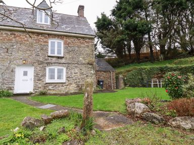 An exterior view of a stone house with white framed windows and door a green lawn with rocks and plants and trees in the background at Lanvean Farmhouse Lanvean near St Mawgan