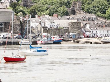 A riverside scene with boats on the water and buildings with stone walls and green trees in the background at Coed Derw Isaf in Betws-Y-Coed
