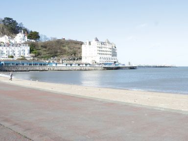 A beach with a promenade a large white hotel on a hillside and a pier extending into the sea