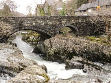 A stone bridge over a river with rocks and buildings in the background at Coed Derw Isaf in Betws-Y-Coed