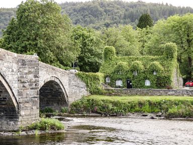 A stone bridge over a river next to a house covered with ivy and trees in the background at Coed Derw Isaf in Betws-Y-Coed