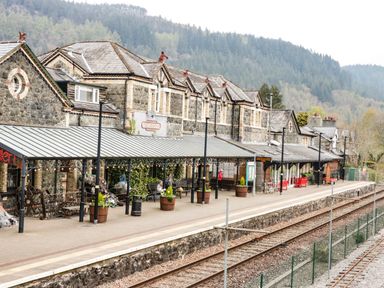 A train station platform with benches and flower pots next to railway tracks and stone buildings at Coed Derw Isaf in Betws-Y-Coed