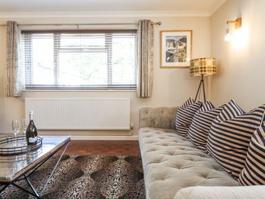 A living room with a large sofa with striped cushions a coffee table with a bottle and glasses a floor lamp and a window at Coed Derw Isaf in Betws-Y-Coed