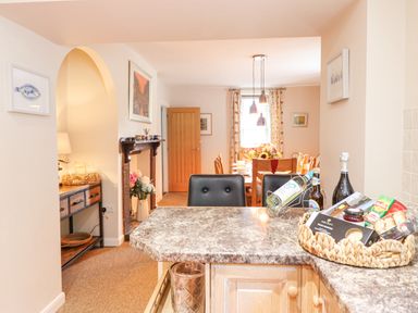 A kitchen island with wine bottles and snacks overlooking a dining area with a wooden table and chairs at Coed Derw Isaf in Betws-Y-Coed