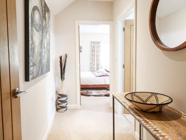 A hallway with a wooden table holding a woven basket a circular wall mirror a door leading to a bedroom with a bed and curtains at Coed Derw Isaf Betws-Y-Coed
