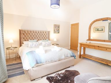 A bedroom with a beige bed and pillows bedside tables and lamps a wooden dressing table with a mirror and a chair at Coed Derw Isaf in Betws-Y-Coed