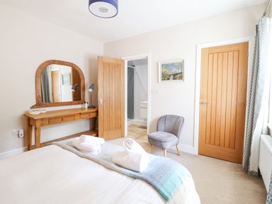 A bedroom with a bed, a wooden dressing table with a mirror, a gray chair, two wooden doors, and a window with curtains at Coed Derw Isaf in Betws-Y-Coed